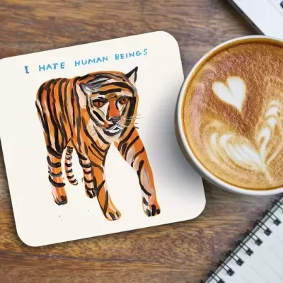 Porta-copos com desenho de tigre e texto 'I HATE HUMAN BEINGS' ao lado de café latte e bloco de notas.