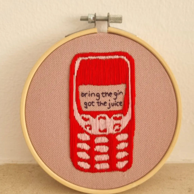 Bordado de telemóvel antigo vermelho e branco com texto no centro em bastidor de madeira sobre tecido cor de rosa