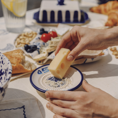 Queijo a ser ralado em prato de cerâmica azul e branca numa mesa com vários alimentos.