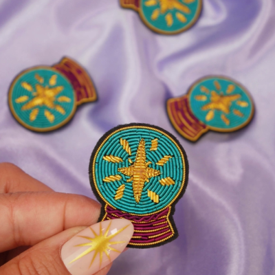 Distintivos bordados em tecido com desenho amarelo sobre círculo azul-turquesa e base vermelha, em fundo lilás.