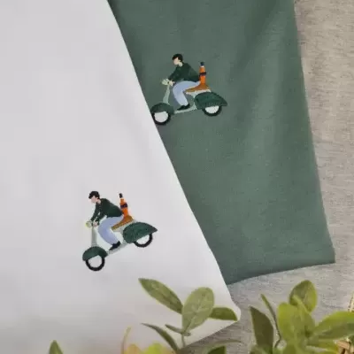 Duas t-shirts com bordado de homem numa scooter, uma branca e outra verde musgo.