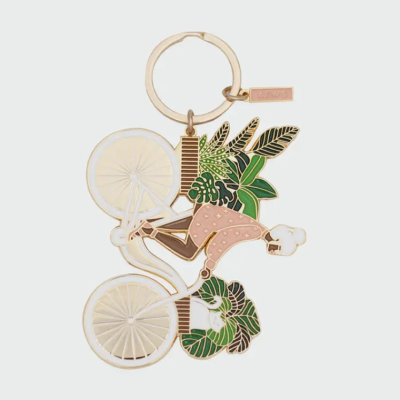 Chaveiro metal dourado com mulher de cabelo branco em bicicleta e folhas verdes.