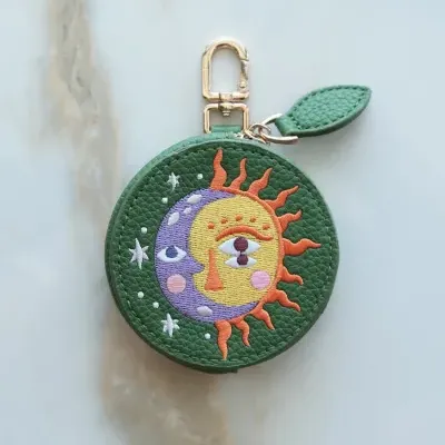Bolsa redonda verde com bordado de sol e lua, fecho dourado e adorno de pele verde, sobre superfície de mármore claro.