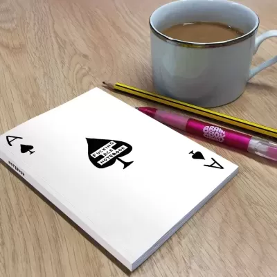 Bloco de notas branco com símbolo de espadas e texto, lápis amarelo e caneta cor-de-rosa perto de chávena branca com chá em mesa de madeira clara
