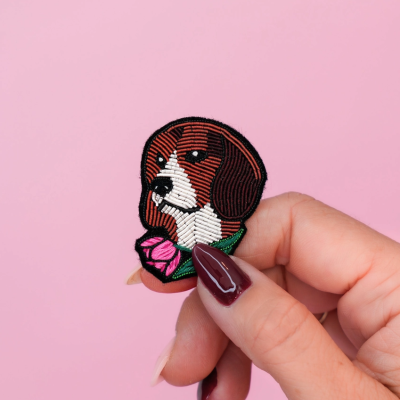 Emblema bordado cão castanho e branco com flor, mãos femininas segurando, fundo rosa.
