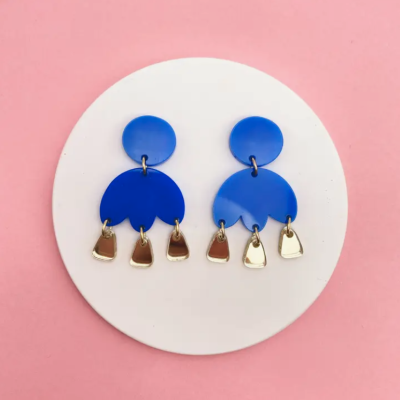 Brincos azuis com pendentes dourados sobre fundo branco circular e rosa
