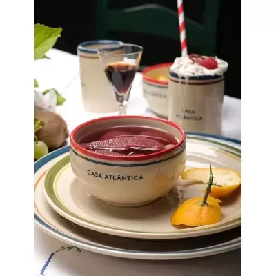 Conjunto de louça em cerâmica bege com bordos vermelhos e linhas coloridas, com tigela, copos e prato sobre mesa clara.