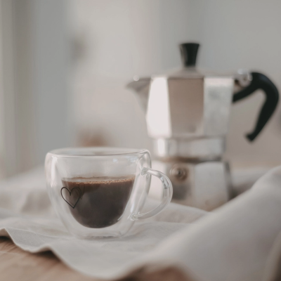 Chávena de café de vidro duplo com desenho de coração e cafeteira de metal ao fundo.