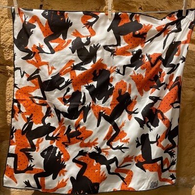 Lenço com padrão de rãs em preto e laranja sobre branco pendurado.