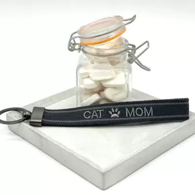 Chaveiro cinza escuro com texto 'CAT MOM' e frasco de vidro com comprimidos brancos em base branca