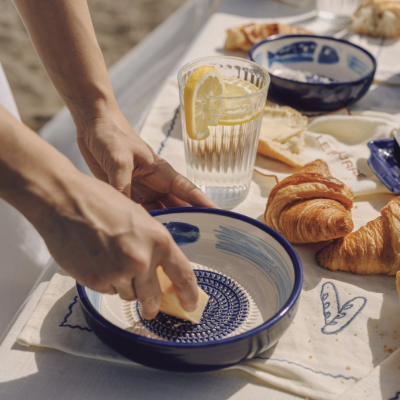 Bowl de cerâmica azul e branco com manteiga, croissants, copo com limão e toalha bordada numa mesa exterior.