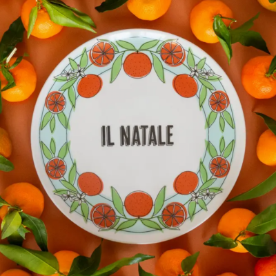 Prato redondo branco com decoração de tangerinas e texto 'IL NATALE', rodeado por tangerinas frescas