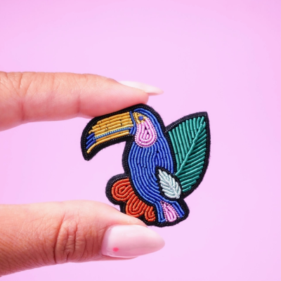 Aplicação bordada colorida de tucano azul com bico amarelo e folhas verdes.