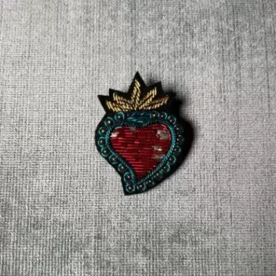 Pin em forma de coração vermelho com contorno azul e detalhes dourados sobre tecido cinzento