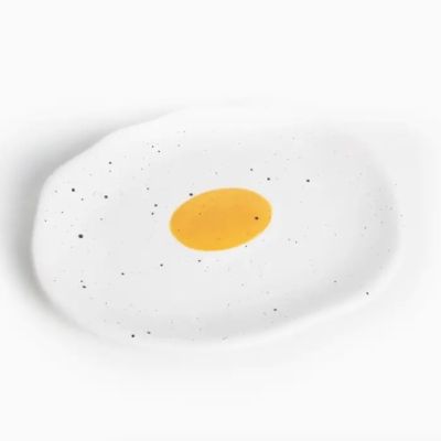 Prato cerâmico branco com padrão de manchas pretas e centro amarelo que simula um ovo estrelado