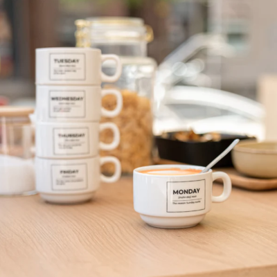 Chávenas de café brancas com nomes dos dias da semana, empilhadas em mesa de madeira clara