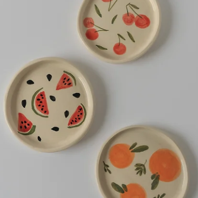 Três pratos cerâmicos pintados com frutas em fundo creme.