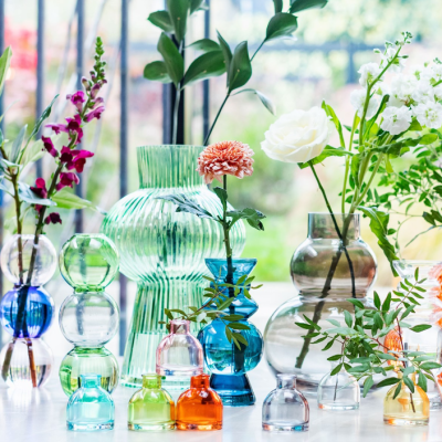 Vasos de vidro colorido com flores em cima de uma mesa branca junto a uma janela