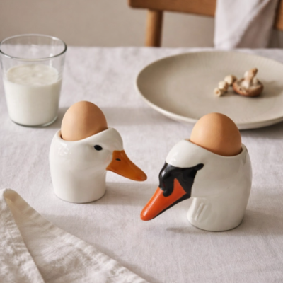Suportes para ovos em forma de cabeça de pato e cisne com ovos castanhos numa mesa branca.