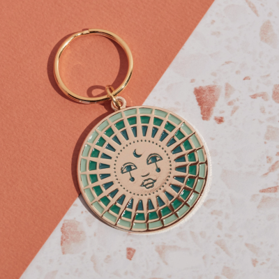 Chaveiro dourado com rosto de sol e padrão verde e azul em fundo bicolor branco e terracota