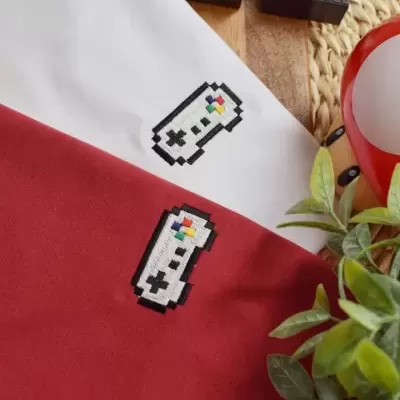 Tecido branco e vermelho com bordados de comandos de videojogo pixelados e decoração pixelada em madeira com planta verde ao lado.
