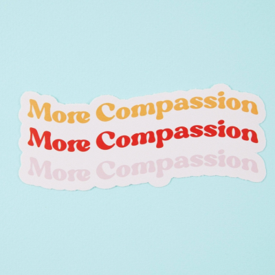 Três autocolantes com o texto 'More Compassion' em laranja, vermelho e rosa claro num fundo azul claro.