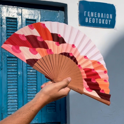 Leque de tecido colorido com estrutura de madeira, mão a segurá-lo, parede branca, janela azul e placa azul com texto em grego