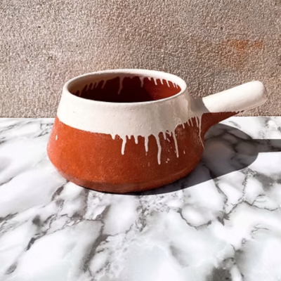 Panela de barro esmaltada branca com castanho avermelhado e alça, sobre mármore.