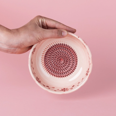 Tigela cerâmica branca e vermelha com padrão em espiral e flores, mão segurando, fundo rosa
