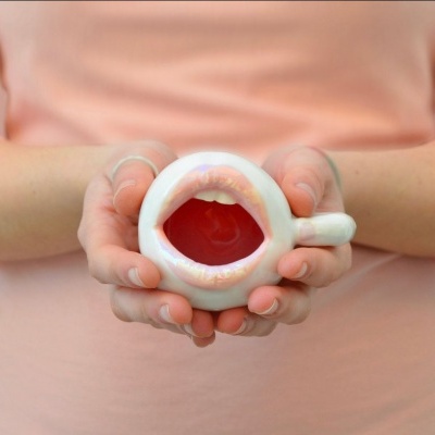 Caneca branca em forma de lábios com interior vermelho, segurada por duas mãos, fundo rosa.