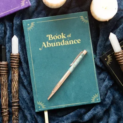 Caderno verde Book of Abundance com caneta, velas e instrumentos com cristais sobre tecido azul escuro