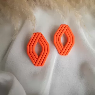 Brincos alaranjados geométricos sobre tecido branco com penas ao fundo.