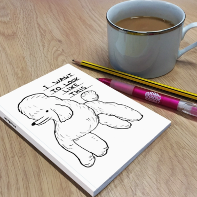 Livro com capa branca com desenho de cão e texto, lápis, caneta rosa e chávena com bebida numa mesa de madeira.