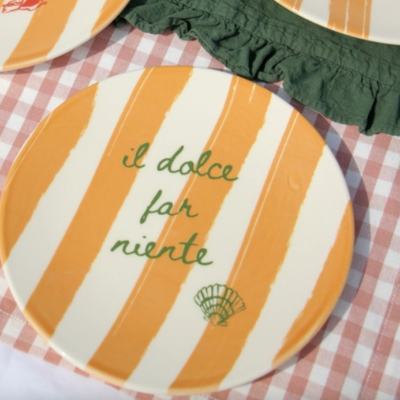 Prato cerâmico listrado laranja e branco com texto em verde e desenho de concha sobre toalha xadrez