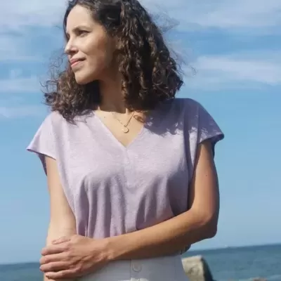 Mulher com t-shirt lilás e saia branca junto ao mar em dia de céu azul