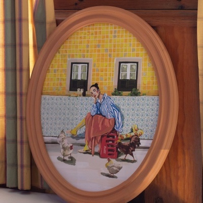 Quadro oval com pintura de mulher e galinhas, fundo de azulejos, contra cortinas às riscas amarelas e verdes.