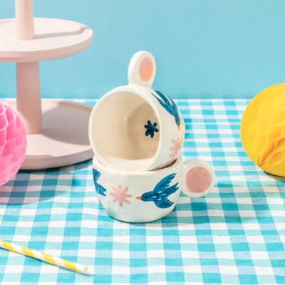 Duas chávenas de cerâmica com decoração de pássaros e flores sobre tecido xadrez azul e branco