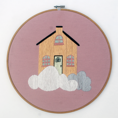 Bordado de casa amarela com nuvens em bastidor de madeira redondo sobre tecido rosa