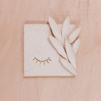 Placa decorativa em cerâmica branca com desenho de olho fechado e folhas em relevo