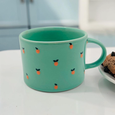 Caneca verde com padrão de pequenas frutas alaranjadas e prato com bolachas.