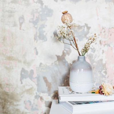 Vaso cerâmico azul e branco com flores secas em cima de livros junto a parede descascada