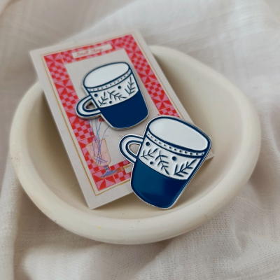 Broches de esmalte em forma de chávenas de café azuis e brancas num cartão decorativo dentro de uma tigela branca.