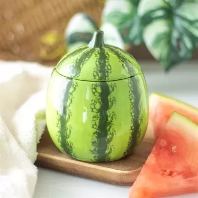 Pote cerâmica forma melancia verde com tampa num tabuleiro de madeira junto a pedaços de melancia e toalha branca