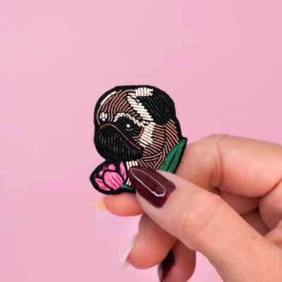 Broche bordado de cão pug castanho e preto com flor, fundo rosa.