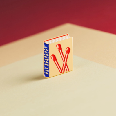 Pin metálico em forma de livro com capa bege e fósforos vermelhos