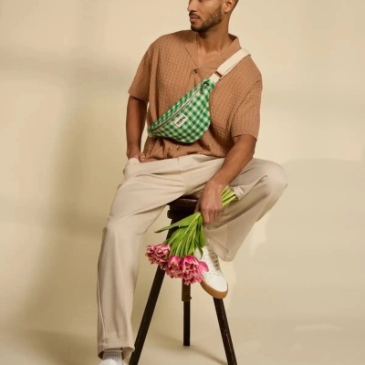 Homem com roupa casual e bolsa verde xadrez sentado num banco a segurar flores cor-de-rosa.