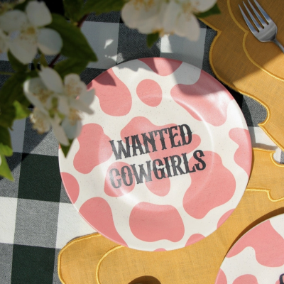 Prato cerâmico rosa e branco com texto 'WANTED COWGIRLS' sobre mesa xadrez verde e branco com guardanapos amarelos e talheres