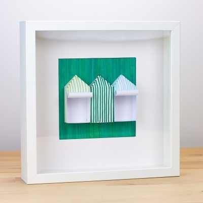 Quadro branco com moldura em madeira contendo três casinhas tridimensionais de papel verde e azul.