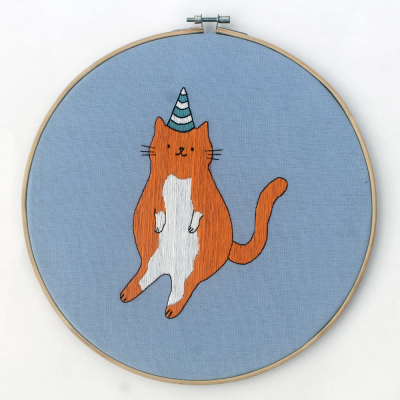 Bordado de gato com chapéu de festa em tecido azul num bastidor de madeira