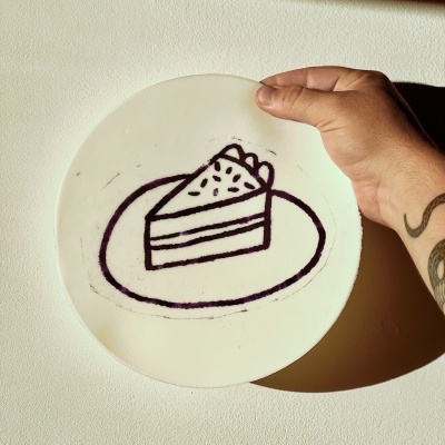Prato branco com desenho preto de fatia de bolo sendo segurado por mão com tatuagem.